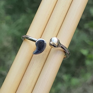 May include: A silver-toned open ring featuring a comma-shaped charm on one end and a small, smooth spherical bead on the other. The band is polished and metallic, designed with an adjustable gap between the two distinct terminal ends.