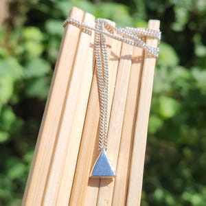 Op de afbeelding: Een zilveren ketting met een driehoekige hanger hangt tegen een achtergrond van lichtbruine bamboestokken. De ketting is delicaat en de hanger heeft een eenvoudige, geometrische vorm. De achtergrond is zachtgroen, wat wijst op een buitenomgeving.