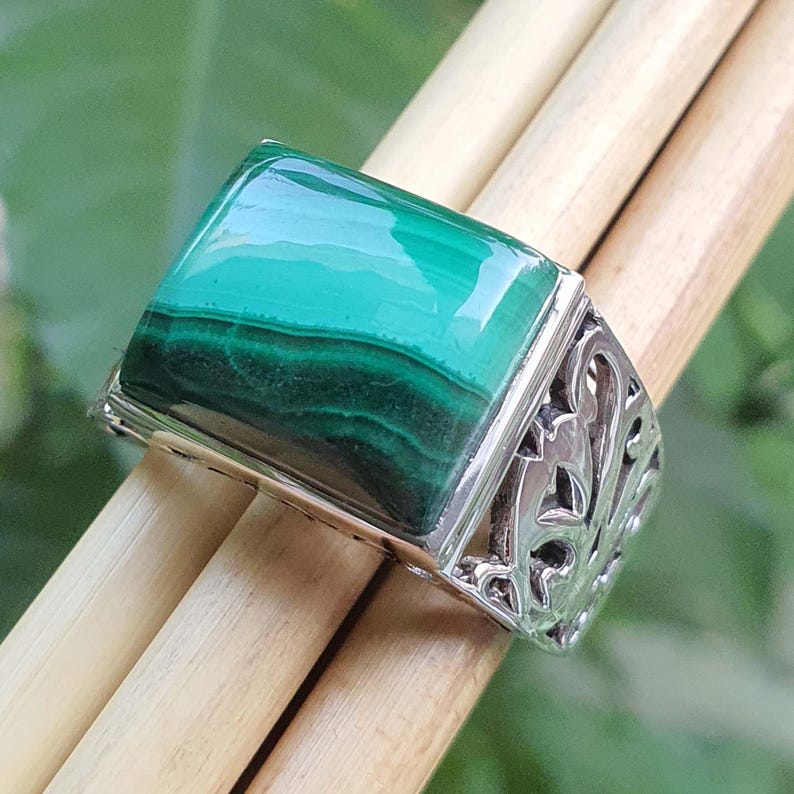 May include: A silver ring featuring a large, rectangular malachite stone with a vibrant green color and dark green banding. The ring has an ornate, openwork design on the sides.