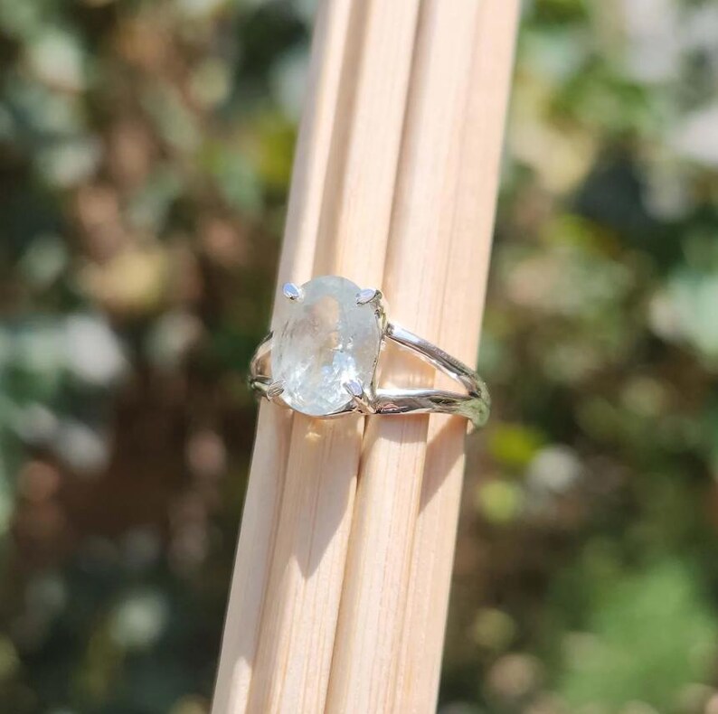 May include: A silver ring featuring a large, oval-shaped, translucent light blue gemstone held in a four-prong setting. The band of the ring has a textured, organic, branch-like appearance with a polished metallic finish.