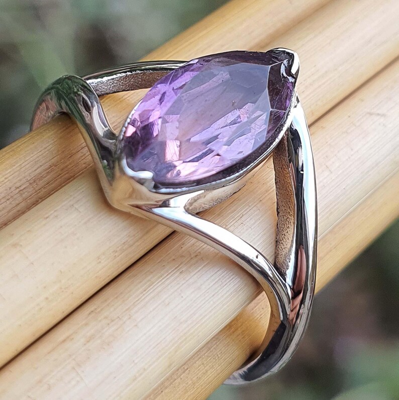 May include: A silver ring featuring a marquise-cut, purple amethyst gemstone. The gemstone is set in a polished silver band with a unique, open design. The ring is displayed on a light brown surface.