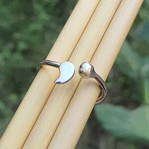 Pode incluir: Um anel aberto de tom prateado com um pendente polido em forma de v&iacute;rgula numa extremidade e uma pequena conta esf&eacute;rica lisa na outra. A banda de metal &eacute; fina e reflexiva, concebida com um espa&ccedil;o ajust&aacute;vel entre as duas extremidades.