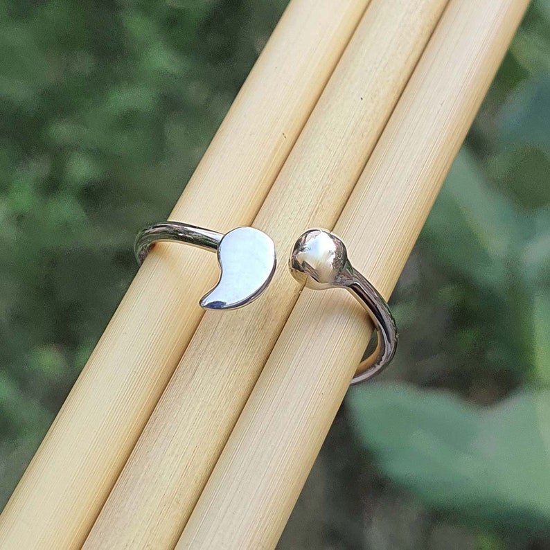 Pode incluir: Um anel aberto de tom prateado com um pendente polido em forma de v&iacute;rgula numa extremidade e uma pequena conta esf&eacute;rica lisa na outra. A banda de metal &eacute; fina e reflexiva, concebida com um espa&ccedil;o ajust&aacute;vel entre as duas extremidades.