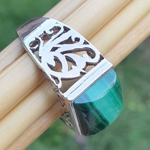 May include: A silver ring with an openwork design and a rectangular, polished green malachite stone. The ring is set on a light brown surface, with a blurred green background.
