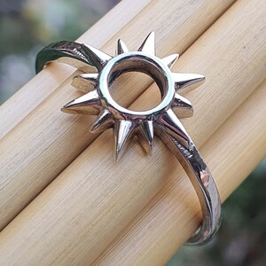 Pode incluir: Um anel de prata com um design de sol. O sol tem um centro redondo e pontos afiados que irradiam do centro.