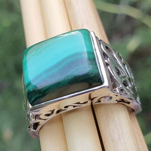 May include: A silver ring featuring a large, rectangular malachite stone. The stone displays bands of green and teal hues. The ring has an ornate, openwork design on the sides, adding detail to the jewelry.