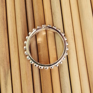 Op de afbeelding: Een zilveren ring met een kralenontwerp. De ring is cirkelvormig en heeft kleine, gelijkmatig verdeelde kralen langs de buitenrand. De ring ligt tegen een achtergrond van lichtbruine houten deuvels.