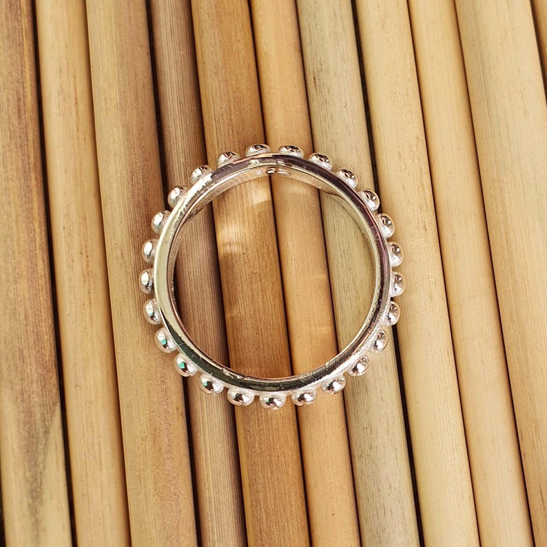 Op de afbeelding: Een zilveren ring met een kralenontwerp. De ring is cirkelvormig en heeft kleine, gelijkmatig verdeelde kralen langs de buitenrand. De ring ligt tegen een achtergrond van lichtbruine houten deuvels.
