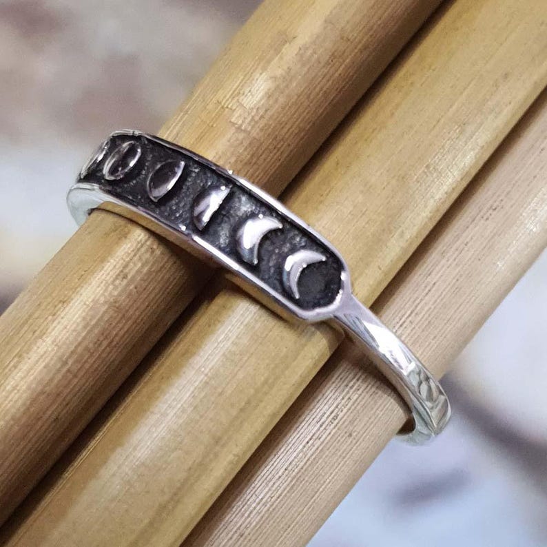 May include: A silver ring featuring a series of moon phases. The ring's band is thin and textured, while the top section displays the moon phases in a dark, contrasting color. The ring is set against a backdrop of wooden dowels.