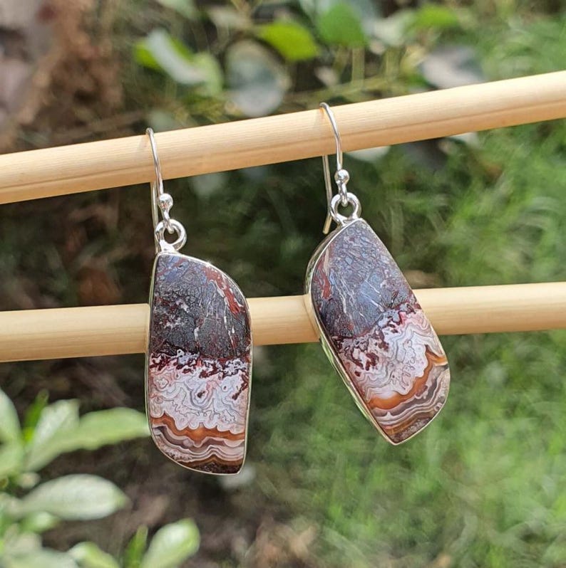 May include: A pair of dangle earrings featuring asymmetrical, polished stones with intricate, wavy banding in shades of dark brown, cream, and rust orange. The stones are set in silver-toned metal frames and hang from simple silver-toned shepherd hook wires.