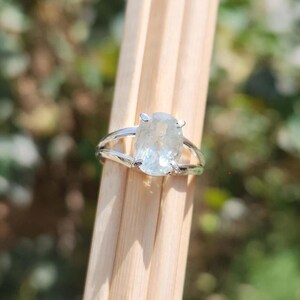 May include: A silver ring featuring a central oval-cut translucent light blue gemstone held by four prongs. The band is split into two thin, polished silver segments that meet at the base of the stone setting, creating a sleek and open design.