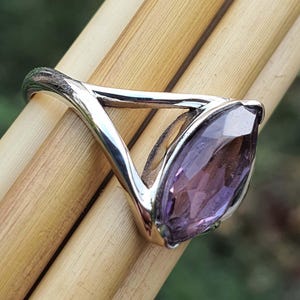 May include: A silver ring with a marquise-cut amethyst gemstone. The ring's band has a unique, open design, and the purple gemstone is set in a bezel setting. The ring is displayed on a light brown surface.