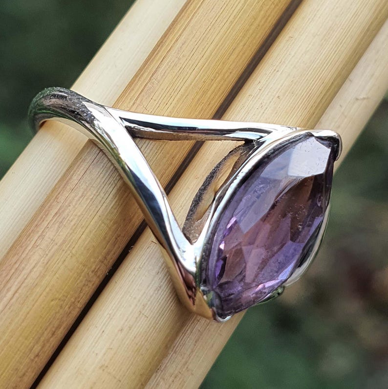 May include: A silver ring with a marquise-cut amethyst gemstone. The ring's band has a unique, open design, and the purple gemstone is set in a bezel setting. The ring is displayed on a light brown surface.