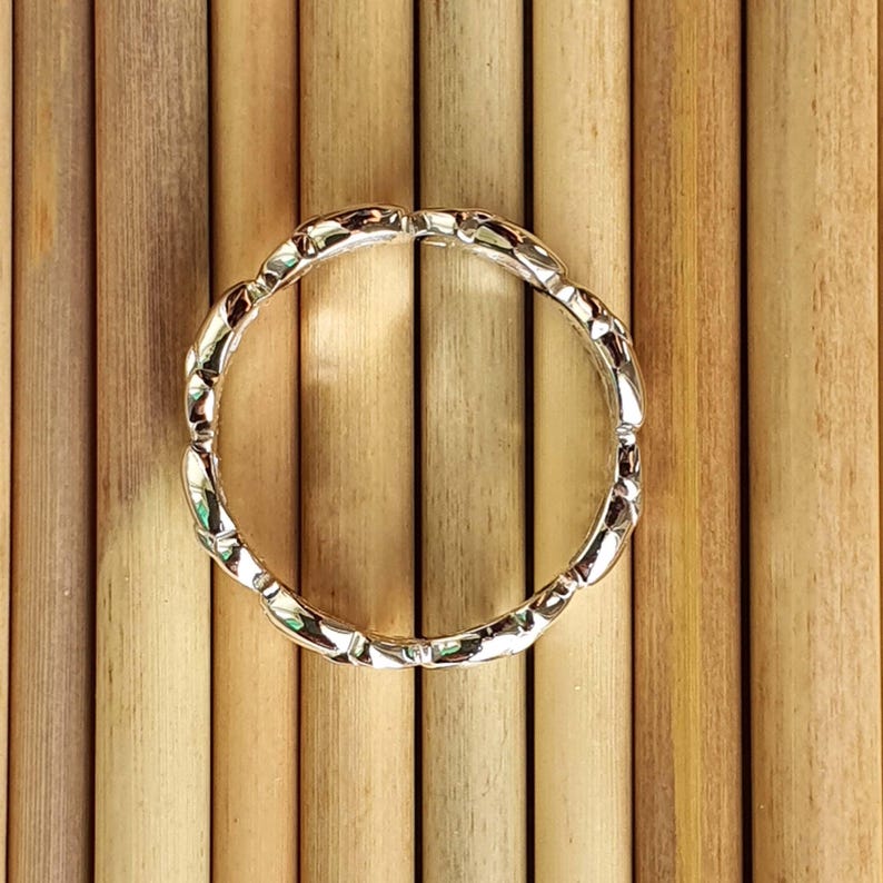 May include: A silver ring with a repeating, textured design. The ring's surface reflects light, creating a shimmering effect. The ring is set against a background of vertical, light brown wooden dowels.