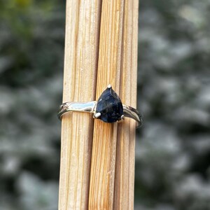 May include: A silver ring with a pear-shaped, dark blue gemstone. The ring is set on a thin band and is displayed against a backdrop of wooden sticks. The gemstone has a pointed top and a smooth, polished surface.