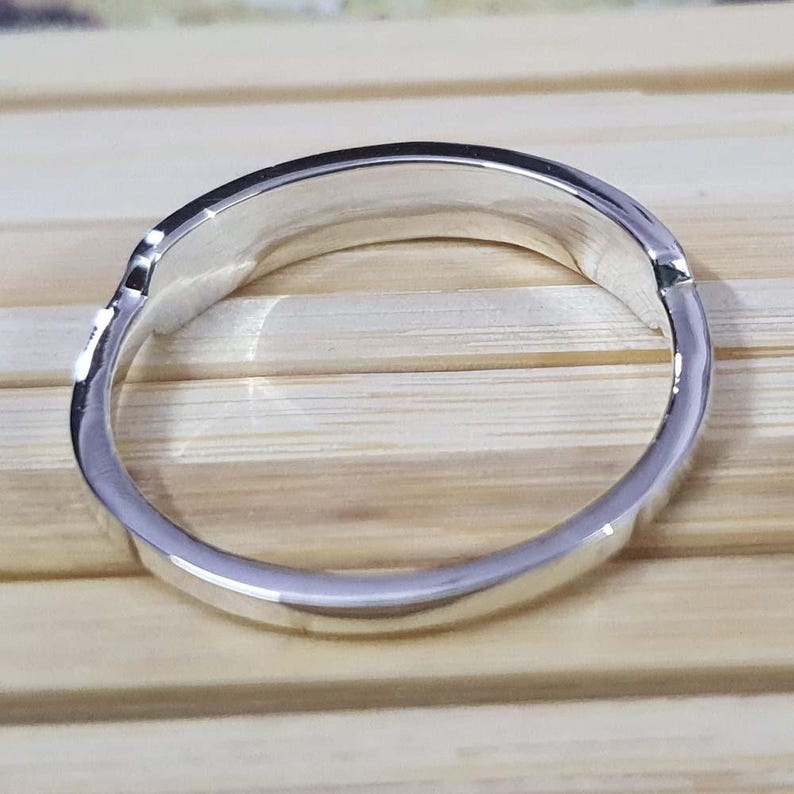 May include: A silver ring with a smooth, polished surface. The ring has a simple, elegant design, reflecting light. The ring is shown on a light brown wooden surface.