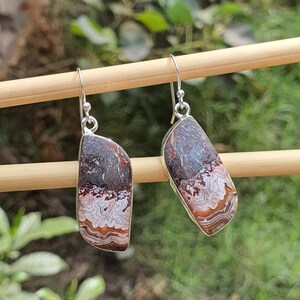 May include: A pair of dangle earrings featuring polished, asymmetrical pieces of Crazy Lace Agate stone. The stones display intricate, swirling bands of white, cream, orange, and deep brown colors, set in silver-toned metal with hook-style ear wires.