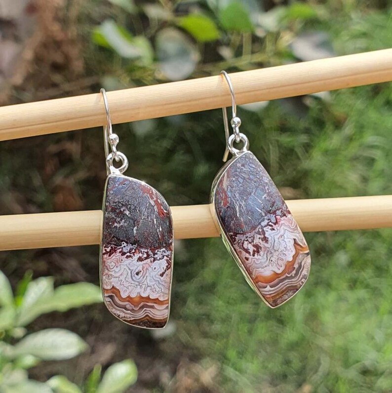 May include: A pair of dangle earrings featuring polished, asymmetrical pieces of Crazy Lace Agate stone. The stones display intricate, swirling bands of white, cream, orange, and deep brown colors, set in silver-toned metal with hook-style ear wires.