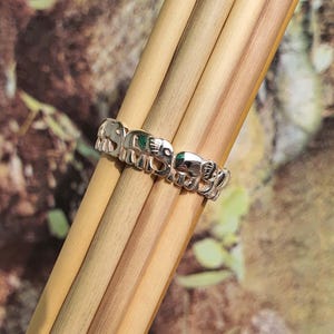 May include: A silver ring featuring a repeating elephant design. The ring is composed of small, detailed elephant figures, with a touch of green. The ring is displayed on a bundle of light brown wooden sticks.