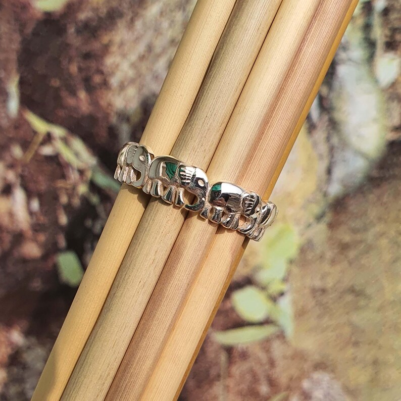 May include: A silver ring featuring a repeating elephant design. The ring is composed of small, detailed elephant figures, with a touch of green. The ring is displayed on a bundle of light brown wooden sticks.