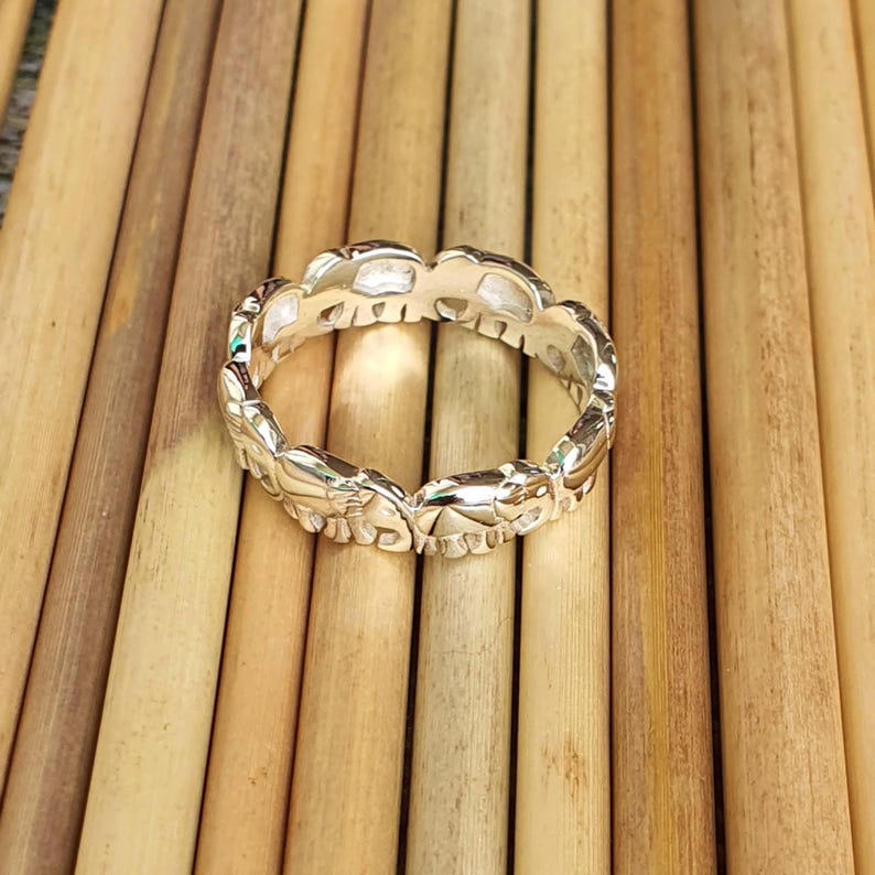 May include: A silver ring featuring a repeating pattern of elephant silhouettes. The ring is made of a shiny, metallic material and is displayed on a light brown, textured surface. The elephants are detailed with trunks, ears, and legs.