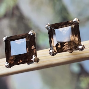 May include: A pair of square-cut smoky quartz stud earrings. Each earring has a dark brown, transparent gemstone set in a silver-coloured metal setting. The earrings are displayed on a light brown surface.