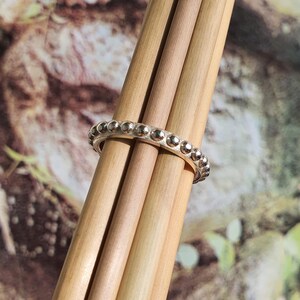 Op de afbeelding: Een zilveren ring met een kralenontwerp. De ring is gewikkeld rond verschillende lichtbruine houten deuvels, met een vage achtergrond van groene en bruine tinten.