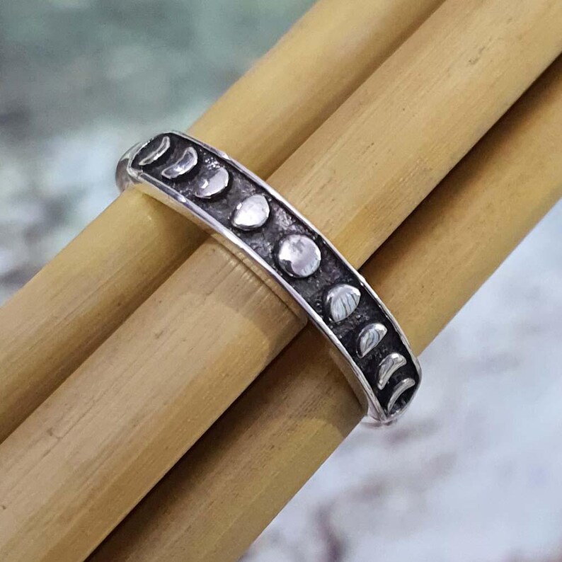 May include: A silver ring featuring a series of moon phase designs. The ring is set against a backdrop of light brown bamboo sticks, creating a contrast between the cool metal and warm wood tones. The ring's design showcases the different phases of the moon.