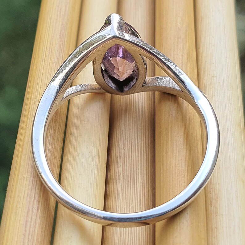 May include: A silver ring with a marquise-cut, faceted, light purple gemstone. The ring's band is a simple, polished silver, and the gemstone is set in a prong setting. The ring is displayed against a light brown, wooden background.