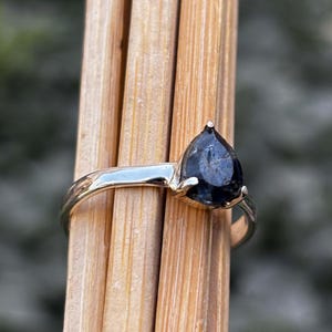 May include: A gold-colored ring featuring a pear-shaped, dark blue gemstone. The gemstone is the focal point, set in a simple, elegant band. The ring is displayed against a backdrop of wooden sticks.