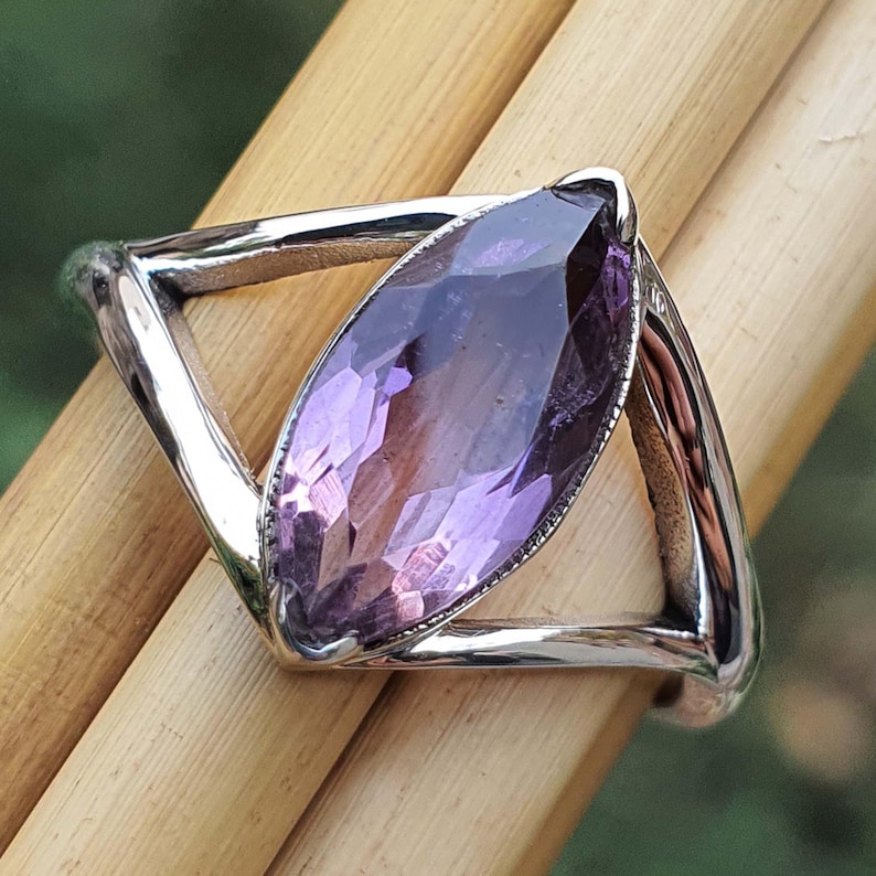 May include: A silver ring featuring a marquise-cut amethyst gemstone. The amethyst is a deep purple color and is set in a geometric, open-work design. The ring is displayed on a wooden surface.