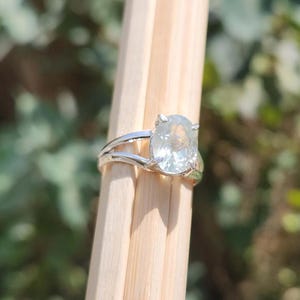 May include: A silver-toned ring featuring a single oval-shaped, translucent white gemstone held in a prong setting. The band is split into two thin, parallel metal strands that meet at the base of the stone, creating a simple and clean design.