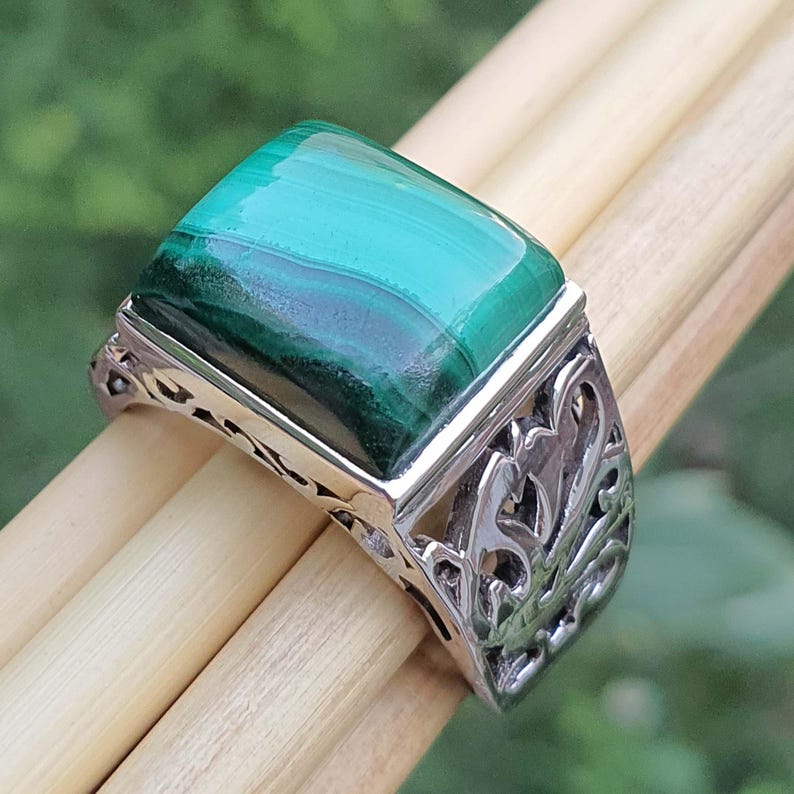 May include: A silver ring with a rectangular malachite stone. The stone is a vibrant green with darker green bands. The ring has an ornate, openwork design on the sides.