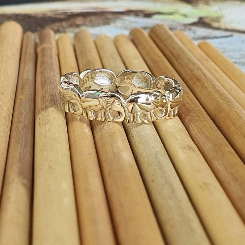 May include: A silver-colored ring featuring a repeating pattern of elephant figures. The ring is set against a background of wooden dowels, creating a contrast of textures and tones. The ring's design showcases the elephants in a detailed, three-dimensional form.