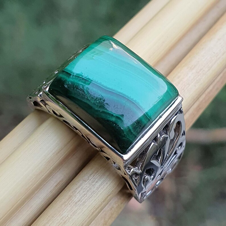 May include: A silver ring with a rectangular malachite stone. The stone is a vibrant green with darker green veining. The ring has an ornate, openwork design on the band.