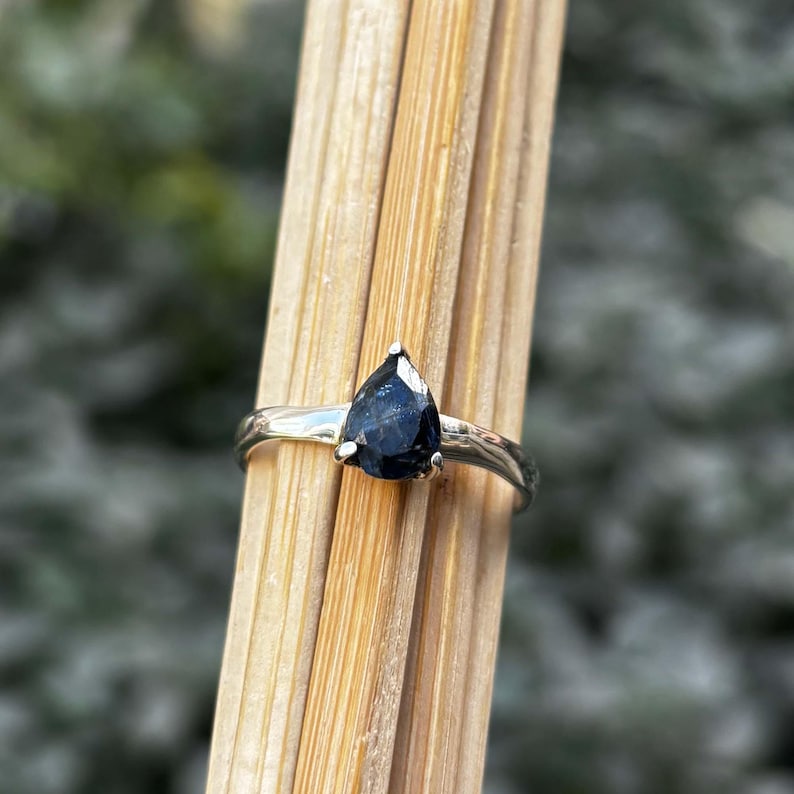 May include: A silver ring featuring a pear-shaped, dark blue gemstone. The ring's band has a simple, elegant design. The gemstone is securely set with small prongs, showcasing its polished surface. The ring is displayed against a backdrop of wooden sticks.