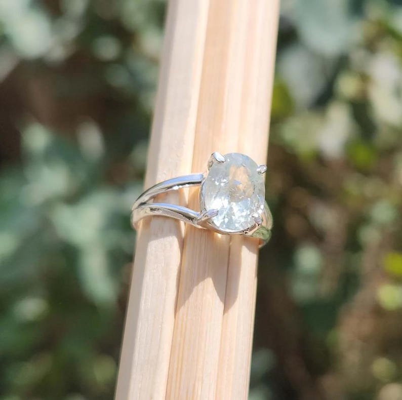 May include: A silver-toned ring featuring a single oval-shaped, translucent white gemstone held in a prong setting. The band is split into two thin, parallel metal strands that meet at the base of the stone, creating a simple and clean design.