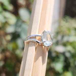 May include: A silver ring featuring a clear, oval-cut gemstone set in a prong setting. The band of the ring is split into two thin, polished silver segments that meet at the base of the stone, creating a sleek and open design for the jewelry piece.