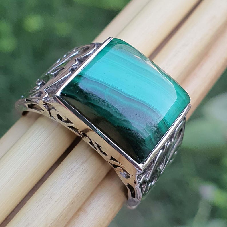 May include: A silver ring with a rectangular malachite stone. The stone is a vibrant green with darker green bands. The ring has an ornate, openwork design on the band. The ring is displayed on wooden dowels.