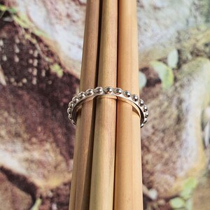 Op de afbeelding: Een zilveren ring met een kralenontwerp. De ring wordt getoond op een bundel lichtbruine houten stokken tegen een vage achtergrond van groene en bruine tinten. Het oppervlak van de ring reflecteert licht, waardoor de kleine, gelijkmatig verdeelde kralen worden benadrukt.