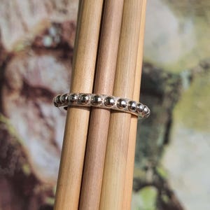 Op de afbeelding: Een zilveren ring met een reeks kleine, ronde, verhoogde kralen die de band omcirkelen. De ring wordt gepresenteerd tegen een achtergrond van verschillende lichtbruine houten deuvels, met een vage achtergrond van groene en bruine tinten.