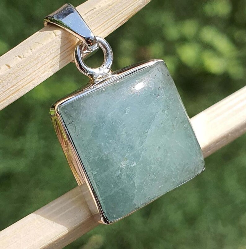 May include: A square-shaped pendant featuring a pale, translucent light green gemstone set in a polished silver-toned metal frame. The pendant includes a circular silver-toned bail at the top, designed for attachment to a necklace chain or cord.