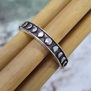 May include: A silver ring featuring a series of moon phase designs. The ring is set against a backdrop of bamboo stalks, highlighting the celestial theme. The ring's design includes crescent, half, and full moon shapes.
