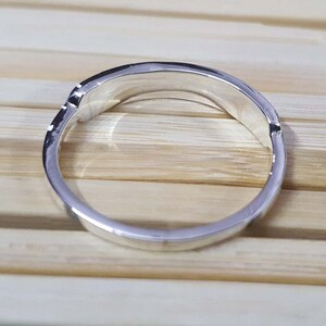 May include: A silver ring with a curved design. The ring is made of a shiny, reflective metal and has a smooth, polished surface. The ring is set against a light brown wooden background.