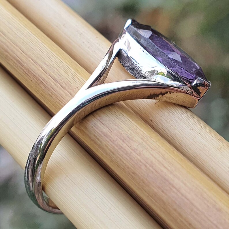 May include: A silver ring with a large, marquise-cut purple gemstone. The ring's band is smooth and polished, and the gemstone is set in a bezel setting. The ring is displayed on a light brown surface.