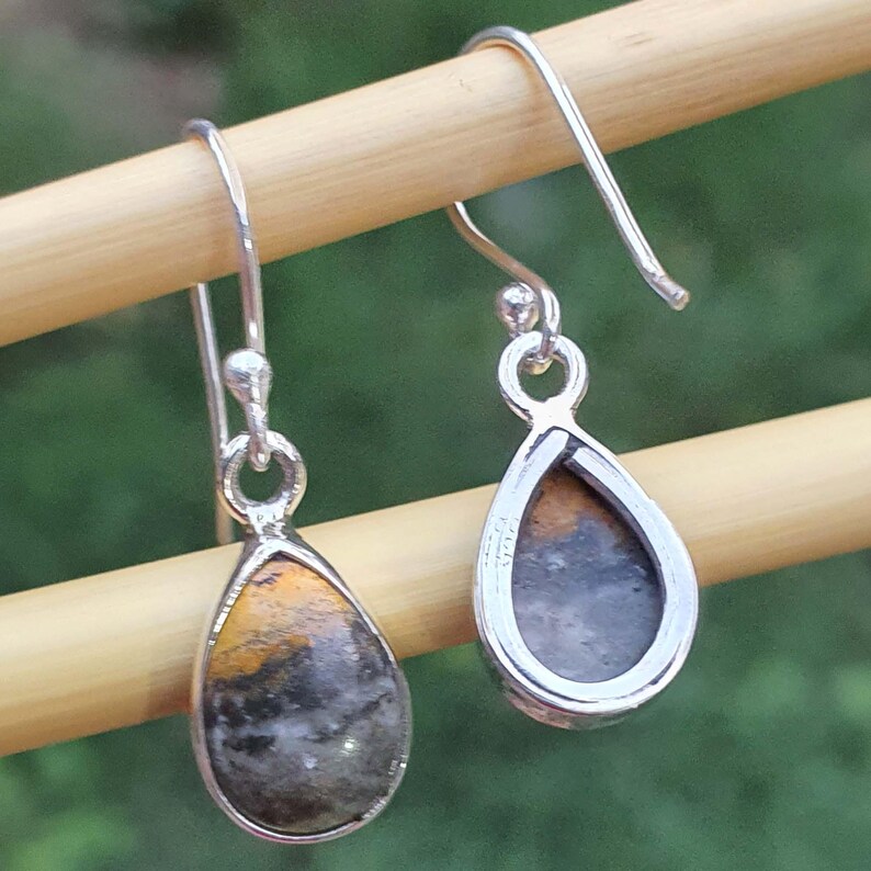 May include: A pair of silver teardrop earrings with a natural stone inlay. The stone features a mix of yellow, black, and gray colors. The earrings are displayed on a wooden dowel against a blurred green background.