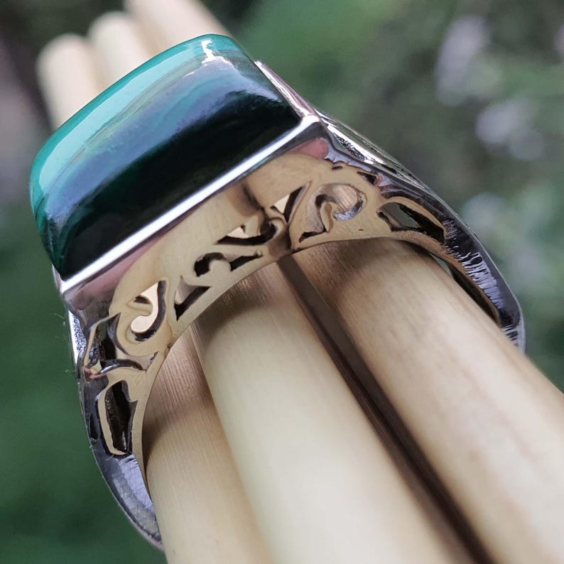 May include: Close-up of a silver ring with a rectangular, green malachite stone. The ring has an ornate, openwork design on the band. The malachite stone has a dark green and teal color.