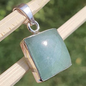 May include: A square-shaped pendant featuring a smooth, pale green gemstone set in a polished silver frame. The pendant includes a silver bail at the top for attachment, showcasing a clean, geometric design with a reflective metallic finish.
