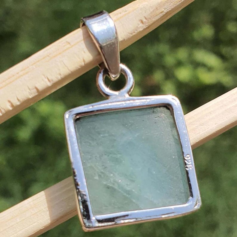 May include: A square pendant featuring a pale green, translucent stone set in a polished silver frame. The metal setting includes a bail at the top for attachment and is clearly stamped with the number 925 on the side of the silver border.