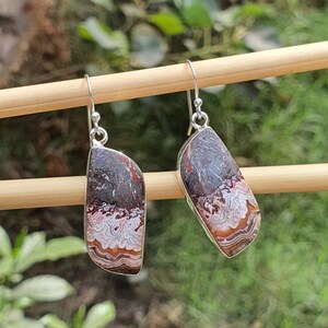May include: A pair of dangle earrings featuring asymmetrical, polished stones with intricate, wavy banding in shades of dark brown, cream, and rust orange. The stones are set in silver-toned metal frames and hang from simple silver-toned shepherd hook wires.